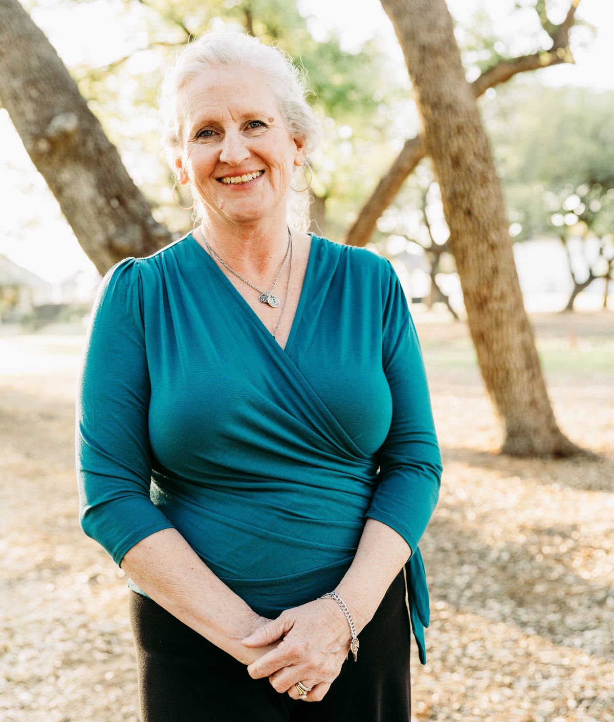 Smiling woman with gray hair wearing a teal blouse standing outdoors among trees.